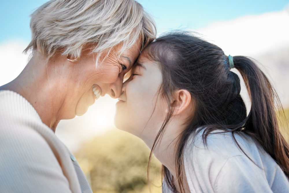 A grandmother and her granddaughter touching foreheads outdoors in the sun