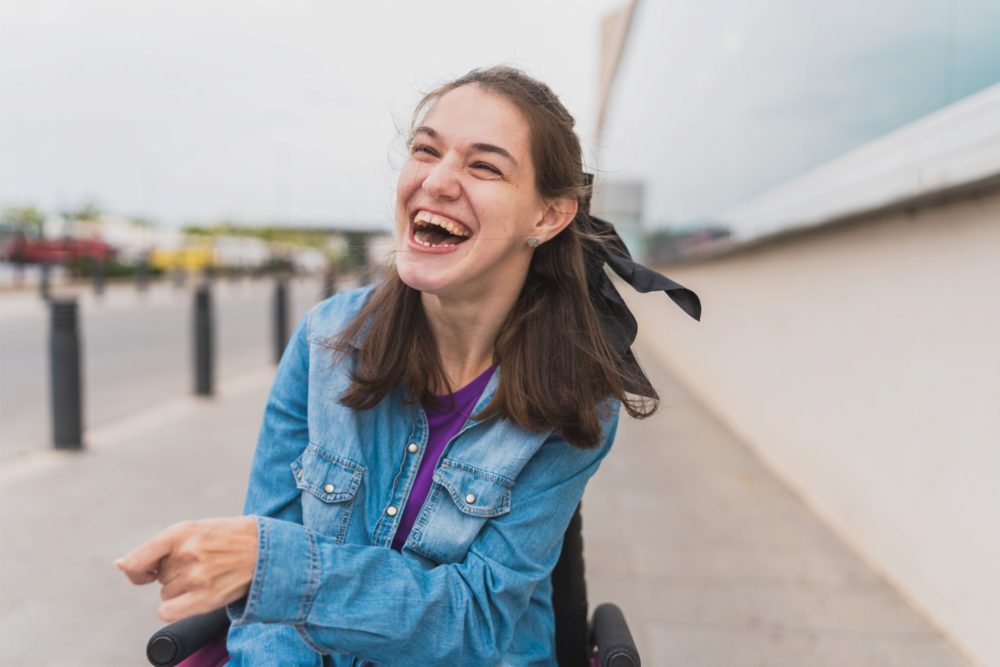 A young woman in a wheelchair laughing outdoors on a city sidewalk